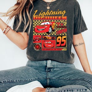 May include: Dark grey t-shirt with a Lightning McQueen design from the Cars film. The graphic includes the text "Lightning McQueen", "95", and "Piston Cup Champion", alongside images of the character.