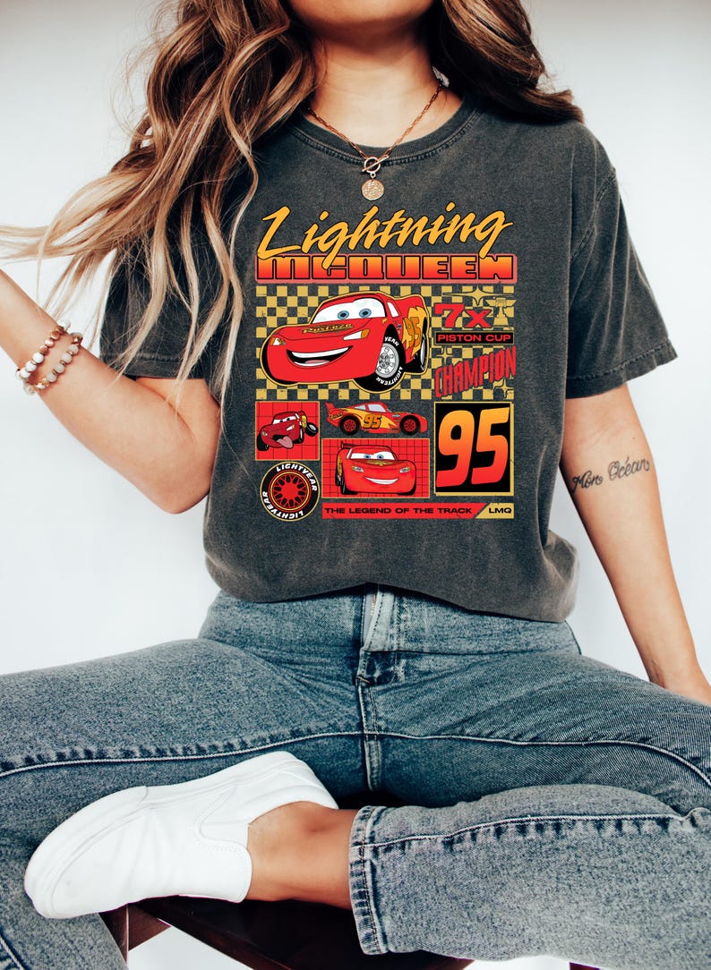 May include: Dark grey t-shirt with a Lightning McQueen design from the Cars film. The graphic includes the text "Lightning McQueen", "95", and "Piston Cup Champion", alongside images of the character.