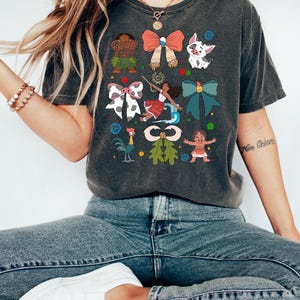 May include: Dark gray t-shirt featuring colorful cartoon characters and bow designs. The shirt's graphic includes a variety of characters and bows in red, white, blue, and green. The shirt is made of a soft material.
