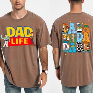 K&ouml;nnte beinhalten: Braunes T-Shirt mit dem Text "DAD LIFE" in Gelb und Blau, mit Illustrationen von Buzz Lightyear und Woody. Auf der R&uuml;ckseite des Shirts steht "DADA DADA" in bunten, cartoonartigen Buchstaben. Ein l&auml;ssiges, bequemes T-Shirt.