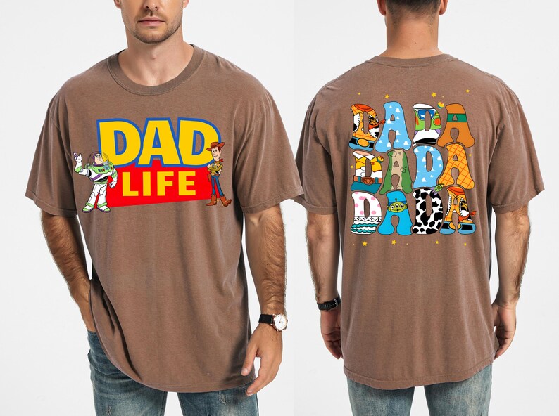 K&ouml;nnte beinhalten: Braunes T-Shirt mit dem Text "DAD LIFE" in Gelb und Blau, mit Illustrationen von Buzz Lightyear und Woody. Auf der R&uuml;ckseite des Shirts steht "DADA DADA" in bunten, cartoonartigen Buchstaben. Ein l&auml;ssiges, bequemes T-Shirt.