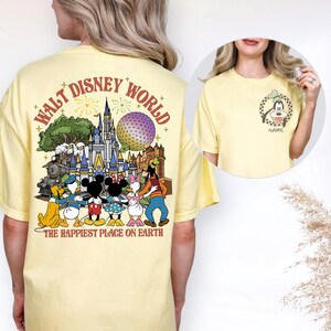 May include: A light yellow t-shirt featuring a colorful graphic of Disney characters and landmarks. The back of the shirt displays the words "WALT DISNEY WORLD" and "THE HAPPIEST PLACE ON EARTH". A smaller image of Goofy is on the front.