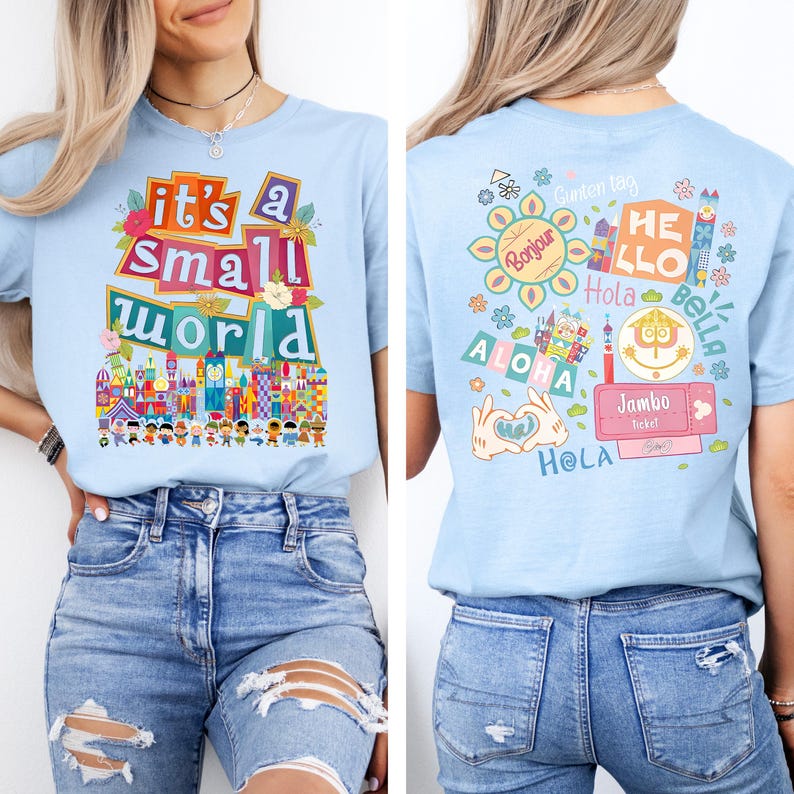 K&ouml;nnte beinhalten: Hellblaues T-Shirt mit dem Text "it's a small world" in bunten Buchstaben und einer Grafik einer skurrilen Stadtlandschaft. Auf der R&uuml;ckseite des Shirts befinden sich verschiedene Gr&uuml;&szlig;e in verschiedenen Sprachen, darunter "Bonjour", "Hola" und "Aloha".