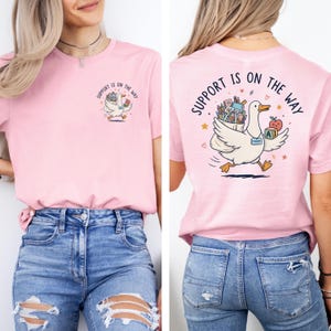 Op de afbeelding: Een lichtroze t-shirt met een kleine afbeelding op de linkerborst van een witte eend die schoolspullen draagt met de tekst "SUPPORT IS ON THE WAY". De achterkant toont een grotere versie van dezelfde eendafbeelding met de tekst.