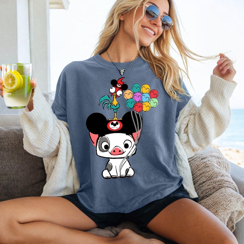 K&ouml;nnte beinhalten: Blaues T-Shirt mit einem Cartoon-Schwein, das einen Mickey-Mouse-Hut tr&auml;gt, mit einem Hahn und bunten Luftballons. Das Shirt hat eine lockere Passform und kurze &Auml;rmel.