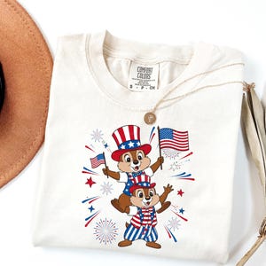 May include: Cream-colored t-shirt featuring a patriotic design with two cartoon chipmunks wearing red, white, and blue outfits and hats, holding American flags. The shirt has the text "COMFORT COLORS" on the tag.
