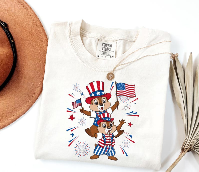May include: Cream-colored t-shirt featuring a patriotic design with two cartoon chipmunks wearing red, white, and blue outfits and hats, holding American flags. The shirt has the text "COMFORT COLORS" on the tag.