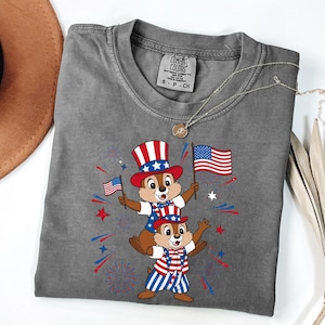 May include: A gray Comfort Colors t-shirt featuring a patriotic design with two cartoon chipmunks wearing red, white, and blue outfits and holding American flags. The design includes fireworks and stars.