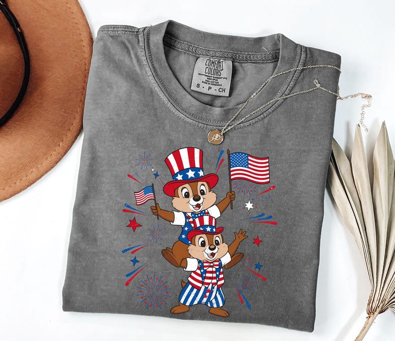 May include: A gray Comfort Colors t-shirt featuring a patriotic design with two cartoon chipmunks wearing red, white, and blue outfits and holding American flags. The design includes fireworks and stars.