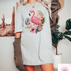 Pu&ograve; includere: T-shirt grigio chiaro con un fenicottero rosa circondato da fiori rosa e foglie verdi. Il fenicottero ha un becco nero ed &egrave; al centro del disegno. La maglietta ha una vestibilit&agrave; ampia e casual.
