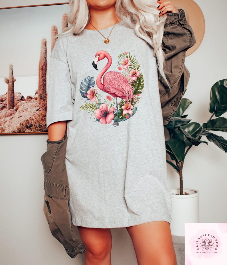 Pu&ograve; includere: T-shirt grigio chiaro con un fenicottero rosa circondato da fiori rosa e foglie verdi. Il fenicottero ha un becco nero ed &egrave; al centro del disegno. La maglietta ha una vestibilit&agrave; ampia e casual.