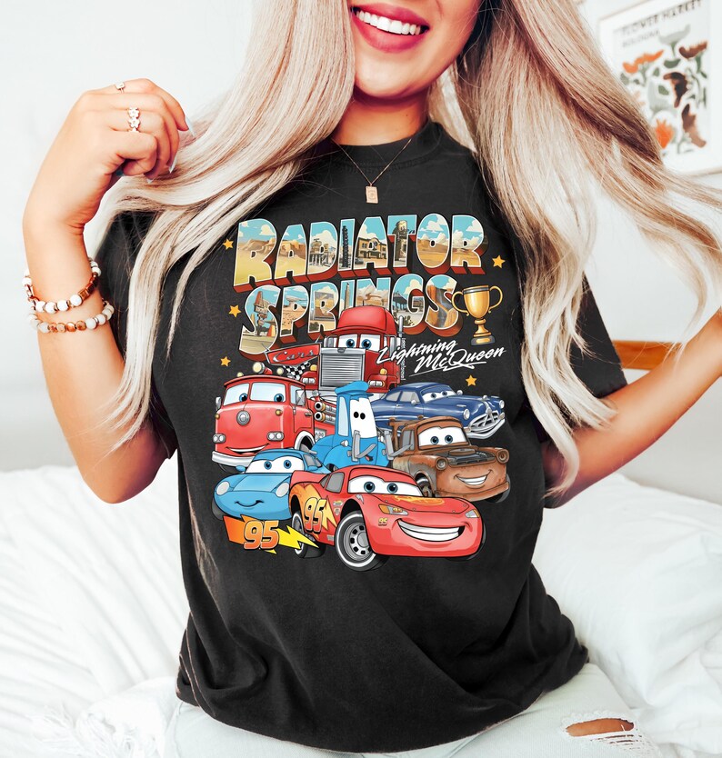 Puede incluir: Camiseta negra con un gr&aacute;fico colorido de personajes de la pel&iacute;cula Cars, incluyendo a Rayo McQueen, y el texto "Radiator Springs". El dise&ntilde;o incluye un trofeo y el n&uacute;mero "95".