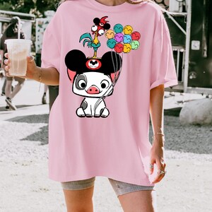 K&ouml;nnte beinhalten: Hellrosa T-Shirt mit einer Cartoon-Schwein-Figur mit Mickey Mouse-Ohren, die bunte Luftballons h&auml;lt. Ein Cartoon-Hahn sitzt auf dem Kopf des Schweins. Das T-Shirt hat eine lockere Passform.
