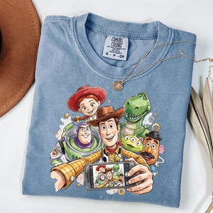 Może przedstawiać: Jasnoniebieska koszulka Comfort Colors z grafiką postaci z Toy Story robiących sobie selfie. Projekt zawiera Woody'ego, Buzz'a Astrala, Jessie, Rexa, Pana Ziemniaka i kosmitę. Złoty naszyjnik jest zarzucony na koszulkę.