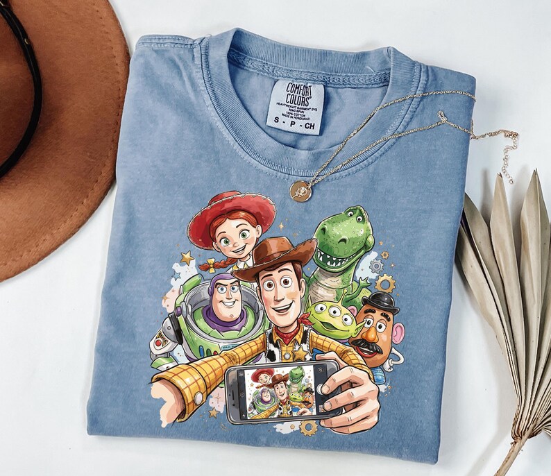 Może przedstawiać: Jasnoniebieska koszulka Comfort Colors z grafiką postaci z Toy Story robiących sobie selfie. Projekt zawiera Woody'ego, Buzz'a Astrala, Jessie, Rexa, Pana Ziemniaka i kosmitę. Złoty naszyjnik jest zarzucony na koszulkę.