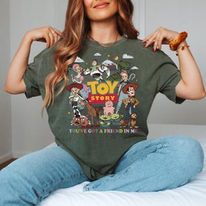 Może przedstawiać: Zielona koszulka z kolorową grafiką postaci z filmu "Toy Story", w tym Woody'ego i Buzza Astrala. Projekt zawiera napisy "TOY STORY" i "YOU'VE GOT A FRIEND IN ME", idealny dla fan&oacute;w filmu.