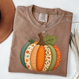 May include: A brown short-sleeve t-shirt featuring a large graphic of a pumpkin made of various patterned fabric segments in shades of orange, green, and cream. The shirt has a crew neckline and a Comfort Colors brand label printed on the inside neck.