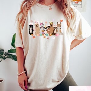 May include: A white t-shirt with a graphic of dogs wearing bunny ears and holding Easter eggs. The dogs are all different breeds and sizes. The graphic is in a cartoon style and is surrounded by flowers.