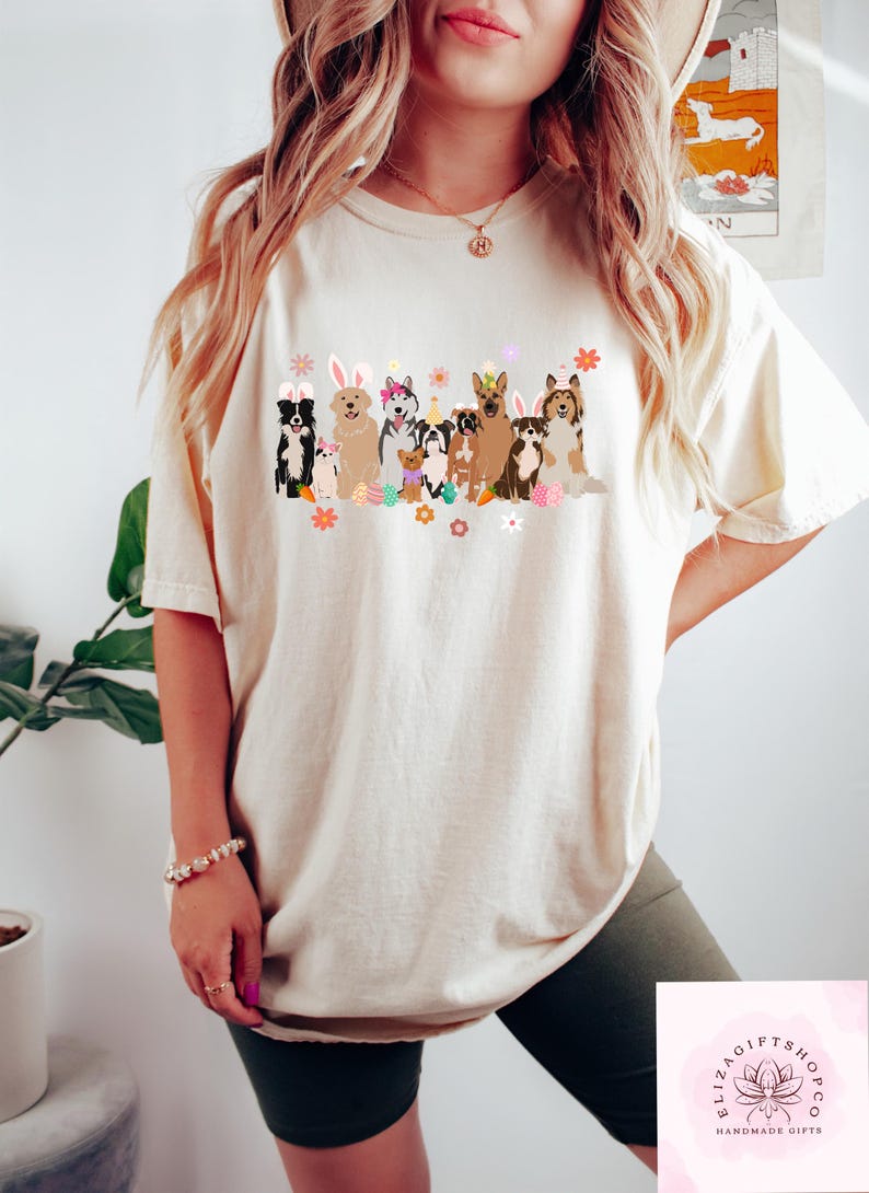 May include: A white t-shirt with a graphic of dogs wearing bunny ears and holding Easter eggs. The dogs are all different breeds and sizes. The graphic is in a cartoon style and is surrounded by flowers.