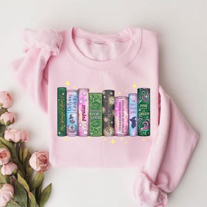 Pu&ograve; includere: Felpa rosa chiaro con una grafica di dorsi di libri colorati, con titoli come "Popular" e "Pink and Green". Il design include accenti di stelle gialle, creando un'estetica stravagante.