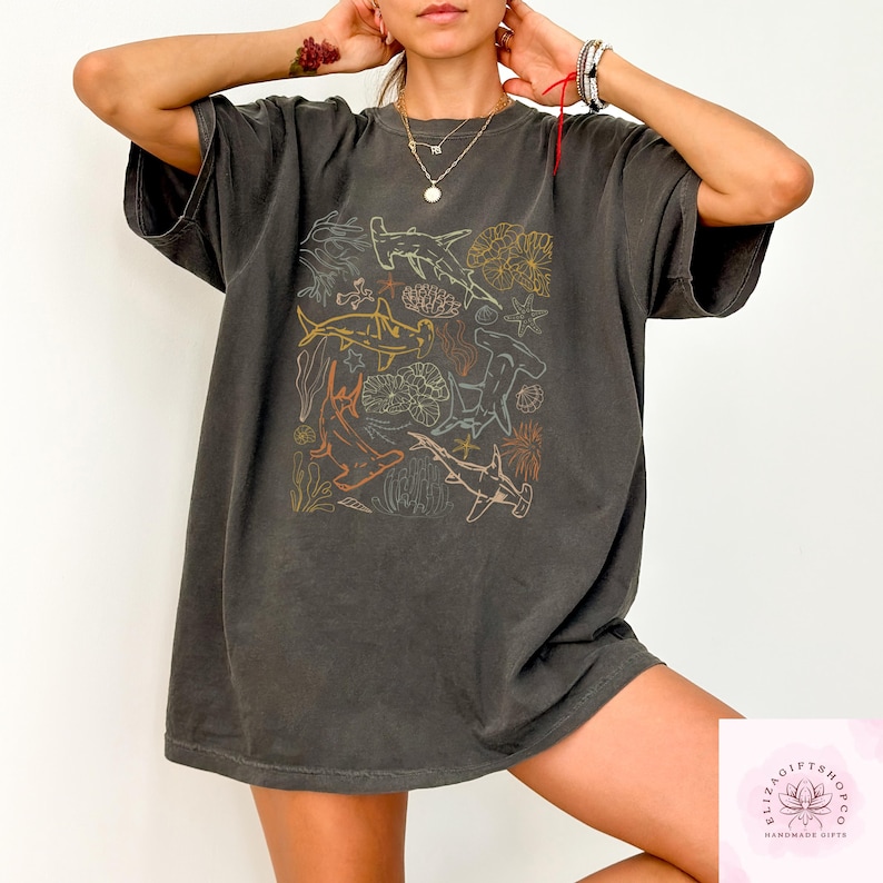 May include: Dark gray t-shirt featuring a colorful illustration of various shark species and coral. The design includes hammerhead sharks, reef fish, and other marine life in orange, yellow, and blue outlines. The shirt has a relaxed fit.