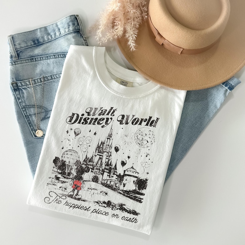 Puede incluir: Camiseta blanca con un gr&aacute;fico negro del castillo de Walt Disney World, Mickey Mouse y el texto "The happiest place on earth". La camiseta se combina con vaqueros azul claro y un sombrero beige.
