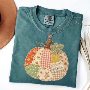 May include: A teal short-sleeved t-shirt featuring a large pumpkin graphic made of various fabric patches including floral, gingham, and lace patterns. The shirt has a Comfort Colors brand label at the neck and a small gold-toned pendant necklace.