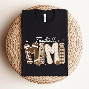 Puede incluir: Una camiseta negra de manga corta con la palabra Football en letra blanca sobre la palabra Mimi en letras grandes. Las letras tienen patrones marrones, de leopardo, de lunares y de cuadros, decoradas con pequeñas estrellas.