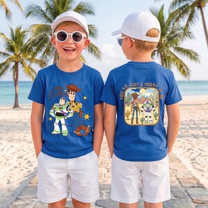May include: A royal blue short-sleeved t-shirt featuring a graphic of Buzz Lightyear and Woody on the front and a desert scene with the text "You've Got A Friend In Me" on the back, paired with white shorts and a white baseball cap.