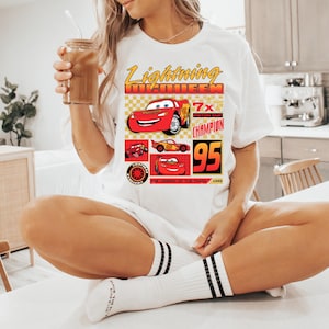 May include: White t-shirt with a graphic of Lightning McQueen from the Cars film. The design includes the text "Lightning McQueen", "7x Piston Cup Champion", and the number "95". The shirt is worn by a person.