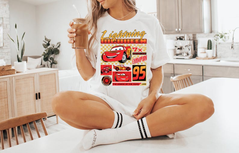 May include: White t-shirt with a graphic of Lightning McQueen from the Cars film. The design includes the text "Lightning McQueen", "7x Piston Cup Champion", and the number "95". The shirt is worn by a person.