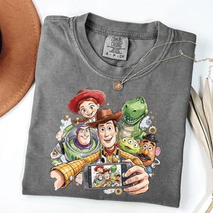 Może przedstawiać: Szara koszulka Comfort Colors z kolorową grafiką postaci z Toy Story robiących sobie selfie. Projekt zawiera Woody'ego, Buzz'a Astrala, Jessie, Rexa, Pana Ziemniaka i kosmitę. Srebrny naszyjnik jest zarzucony na koszulkę.