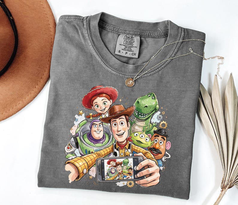 Może przedstawiać: Szara koszulka Comfort Colors z kolorową grafiką postaci z Toy Story robiących sobie selfie. Projekt zawiera Woody'ego, Buzz'a Astrala, Jessie, Rexa, Pana Ziemniaka i kosmitę. Srebrny naszyjnik jest zarzucony na koszulkę.