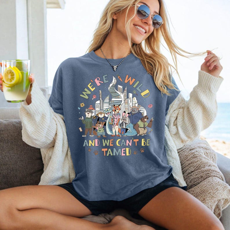 Puede incluir: Una camiseta azul con el texto "WE'RE WILD AND WE CAN'T BE TAMED" y un gr&aacute;fico colorido de personajes animales frente a un horizonte urbano. La camiseta es llevada.