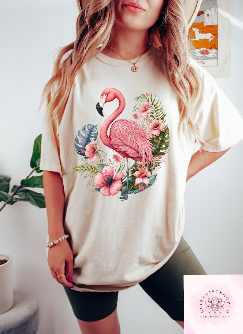 Pu&ograve; includere: T-shirt color crema con illustrazione di un fenicottero rosa circondato da fiori rosa e foglie verdi. Il fenicottero ha un becco nero. La maglietta &egrave; in stile casual.