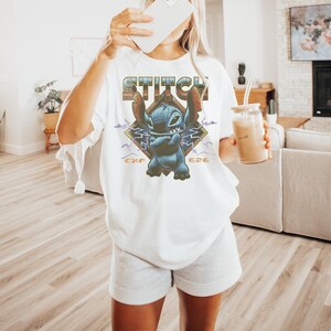 Może przedstawiać: Biała koszulka z grafiką Stitcha z Lilo & Stitch. Projekt zawiera napis "Stitch", wizerunek Stitcha oraz numery "626" i "CKE". Koszulka ma luźny, obszerny kr&oacute;j.