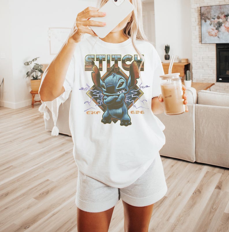 Może przedstawiać: Biała koszulka z grafiką Stitcha z Lilo & Stitch. Projekt zawiera napis "Stitch", wizerunek Stitcha oraz numery "626" i "CKE". Koszulka ma luźny, obszerny kr&oacute;j.