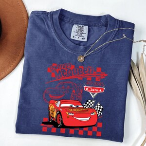 May include: A navy blue t-shirt with a graphic of Lightning McQueen from the Cars film. The design includes the text "Lightning McQueen EST 2006 Cars" in red, with checkered flags. A gold necklace with a pendant is draped across the shirt.
