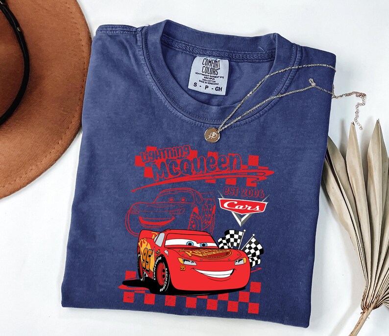 May include: A navy blue t-shirt with a graphic of Lightning McQueen from the Cars film. The design includes the text "Lightning McQueen EST 2006 Cars" in red, with checkered flags. A gold necklace with a pendant is draped across the shirt.