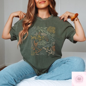 May include: An olive green t-shirt featuring a marine life design. The graphic includes various shark species, coral, and other sea elements in shades of yellow, orange, and white. The shirt is made of a soft material and is suitable for casual wear.
