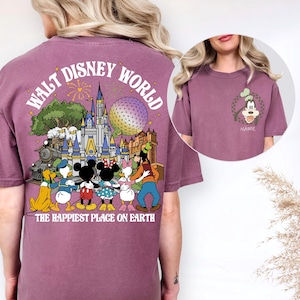 May include: Mauve t-shirt featuring a colorful graphic of Disney World characters and landmarks. The back of the shirt displays the words "Walt Disney World" and "The Happiest Place on Earth". A smaller image of Goofy is on the front.