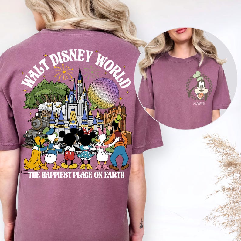 May include: Mauve t-shirt featuring a colorful graphic of Disney World characters and landmarks. The back of the shirt displays the words "Walt Disney World" and "The Happiest Place on Earth". A smaller image of Goofy is on the front.