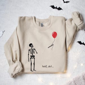 May include: A beige crewneck sweatshirt featuring a black graphic of a skeleton looking at a detached skeletal arm holding a red balloon. The text "Well, shit..." is printed in black below the graphic. The sweatshirt has long sleeves and ribbed cuffs.