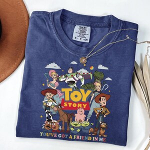 Może przedstawiać: Granatowa koszulka z kolorową grafiką postaci z filmu animowanego "Toy Story". Projekt zawiera Woody'ego, Buzza Astrala i inne postacie, z tekstem "TOY STORY" i "YOU'VE GOT A FRIEND IN ME". Srebrny naszyjnik spoczywa na koszulce.