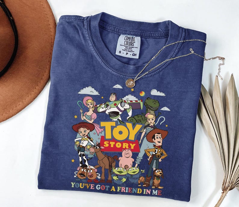 Może przedstawiać: Granatowa koszulka z kolorową grafiką postaci z filmu animowanego "Toy Story". Projekt zawiera Woody'ego, Buzza Astrala i inne postacie, z tekstem "TOY STORY" i "YOU'VE GOT A FRIEND IN ME". Srebrny naszyjnik spoczywa na koszulce.