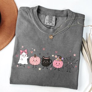 May include: A charcoal gray short-sleeve t-shirt featuring a graphic print of a white ghost with a pink bow, a pink pumpkin, a black cat mug, a pink pumpkin bucket filled with candy, and a spider web, all with small pink stars and hearts.