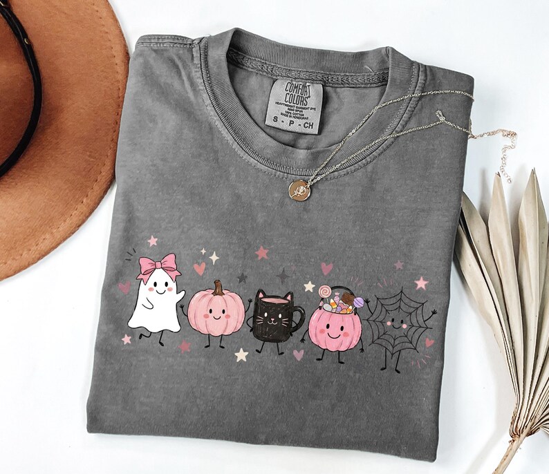 May include: A charcoal gray short-sleeve t-shirt featuring a graphic print of a white ghost with a pink bow, a pink pumpkin, a black cat mug, a pink pumpkin bucket filled with candy, and a spider web, all with small pink stars and hearts.