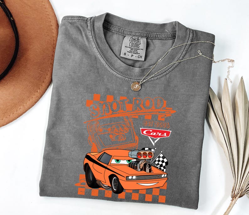 May include: Grey t-shirt with an orange cartoon car design, featuring the text "Shoot Rod" and "Cars". The design includes checkered flags and a vintage car illustration. The shirt is a Comfort Colors brand.