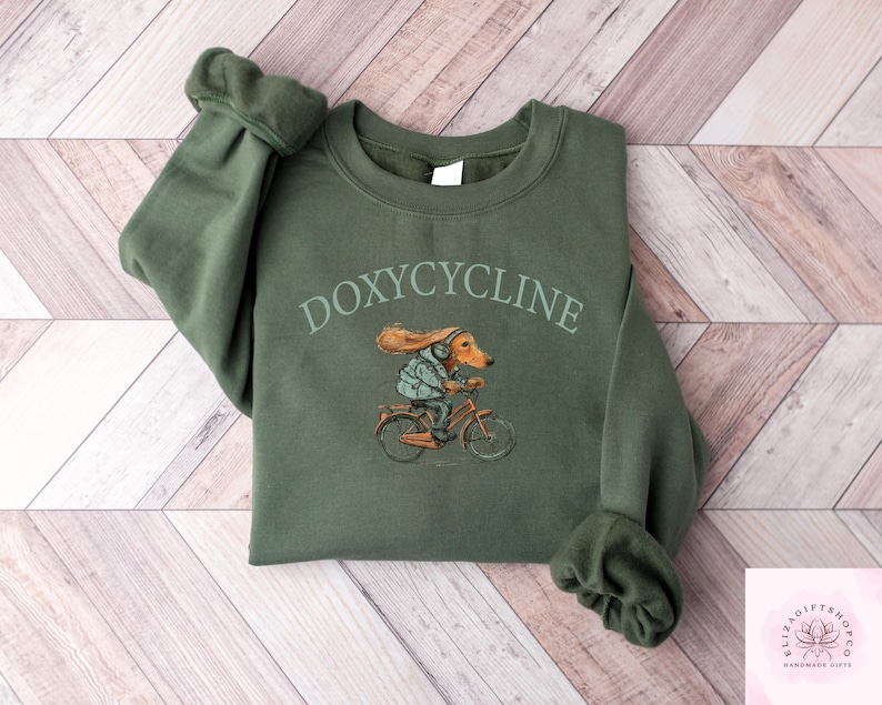 May include: An olive green sweatshirt with the word "DOXYCYCLINE" arched across the top. Below the text is a whimsical illustration of a dachshund riding a bicycle. The dog is wearing a jacket and helmet.