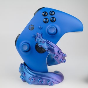 May include: A blue video game controller resting on a stand with a tentacle design. The controller has black buttons and the stand is a gradient of blue and purple. The stand is designed to hold the controller upright.